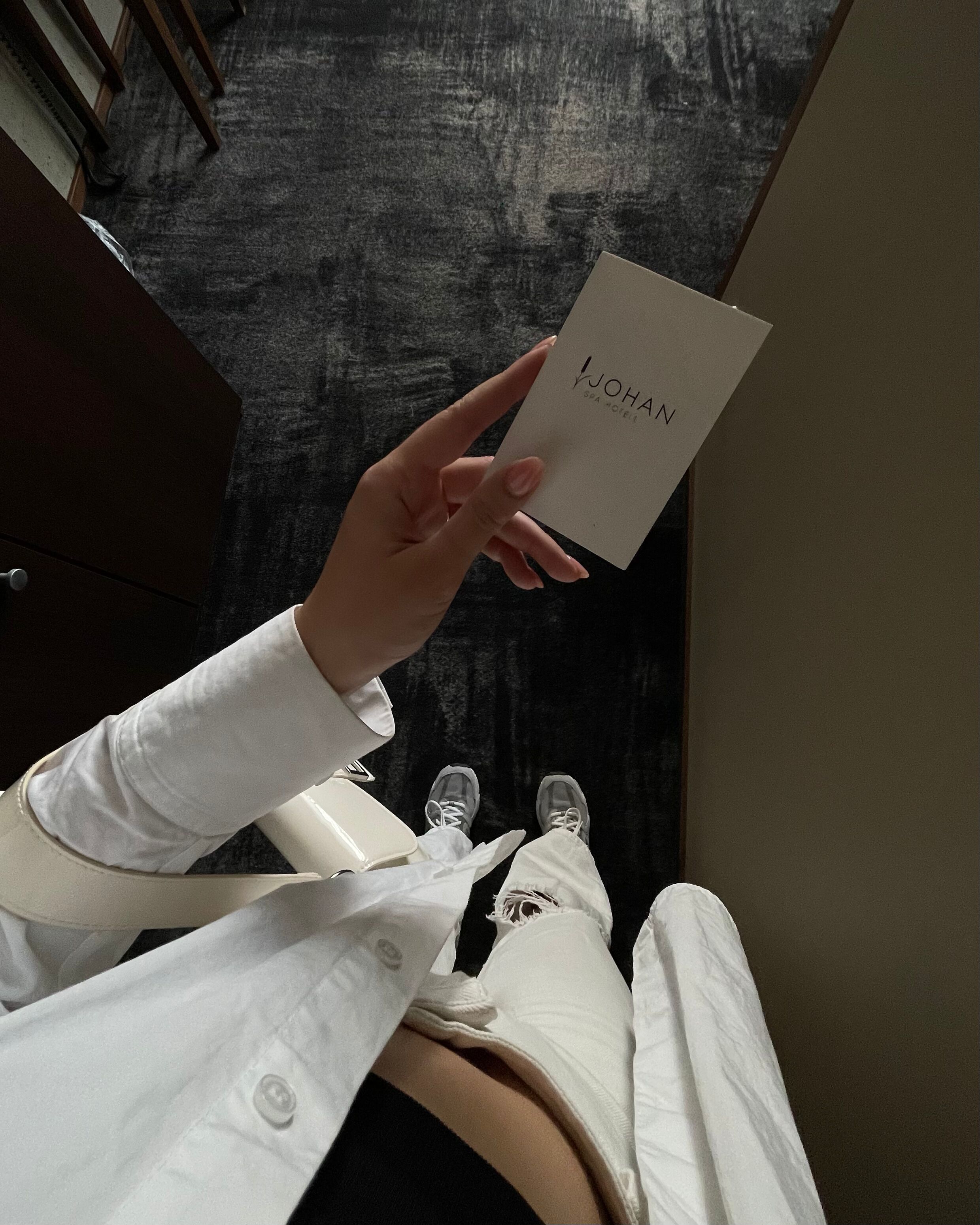 A person holding a card with "JOHAN" visible, standing on a textured floor with a dark wall in the background.