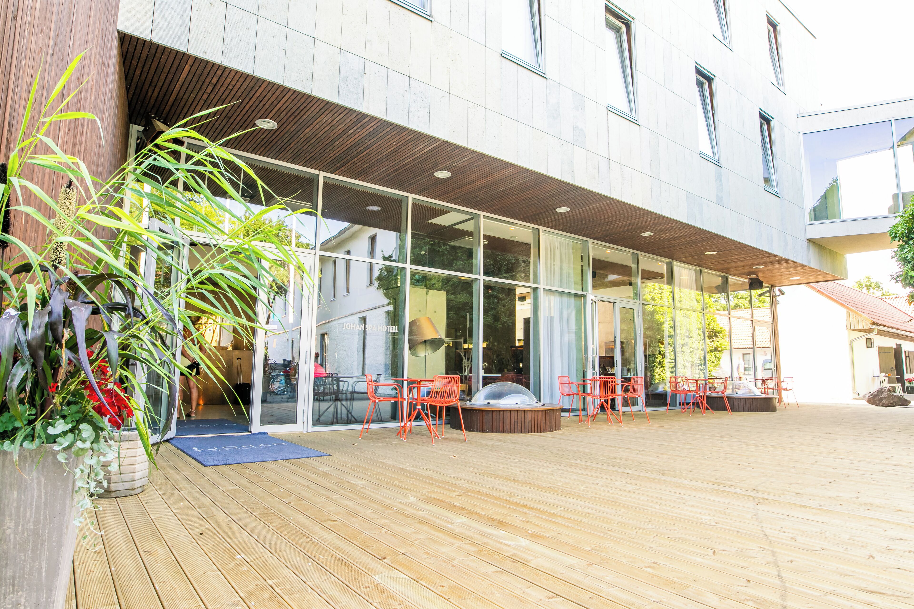 Un edificio moderno con grandes ventanales, terraza de madera y zona de estar al aire libre con sillas naranjas.