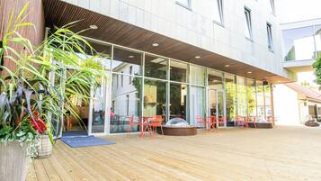 Un edificio moderno con grandes ventanales, terraza de madera y zona de estar al aire libre con sillas naranjas.