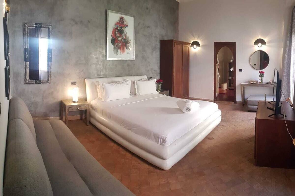 A bedroom with a large bed, a nightstand with a lamp, a wooden wardrobe, a desk with a computer, and a wall-mounted mirror.