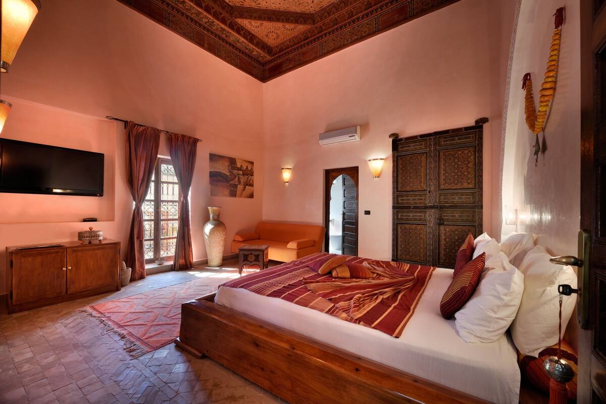 A bedroom with a large bed, wooden headboard, a flat-screen TV, a wooden cabinet, and a decorative wall hanging.