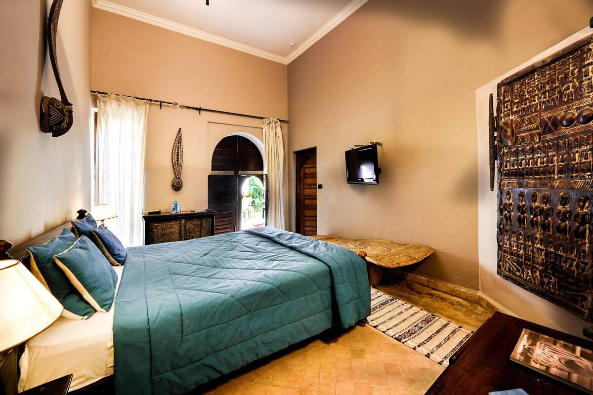A bedroom with a bed, a nightstand, a lamp, a window with curtains, a wooden door, and a decorative wall piece.