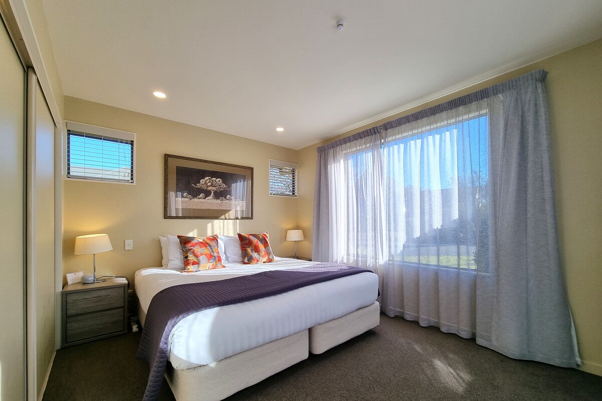 A hotel room with a large bed, a nightstand, a lamp, a framed picture on the wall, and a window with curtains.