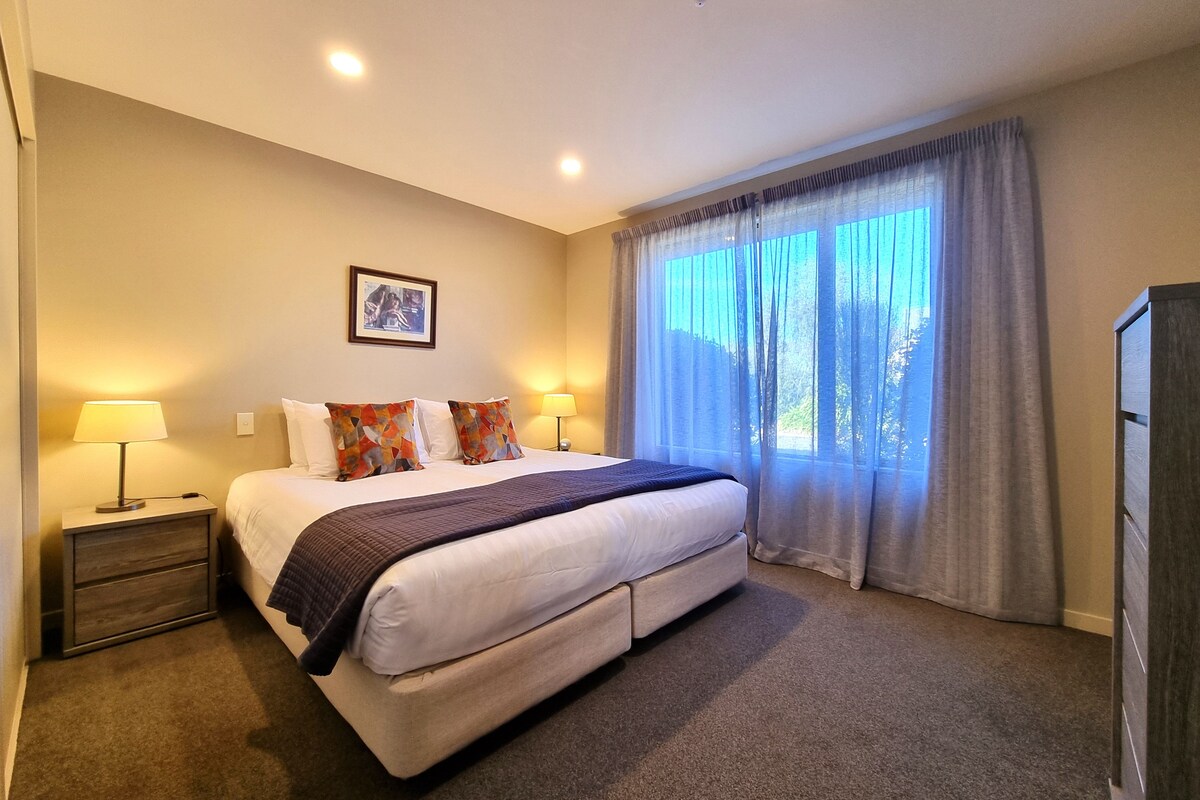 A hotel room with a bed, two bedside lamps, a picture on the wall, and a window with curtains.