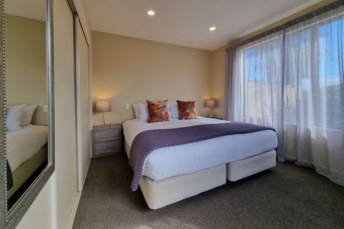 A bedroom with a large bed, two bedside tables, a mirror, and a window with sheer curtains.