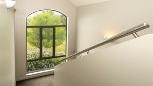 A staircase with a metal handrail leading to a landing, a large arched window with a view of greenery, and a light fixture on the wall. - Vintner's Retreat (Rapaura)