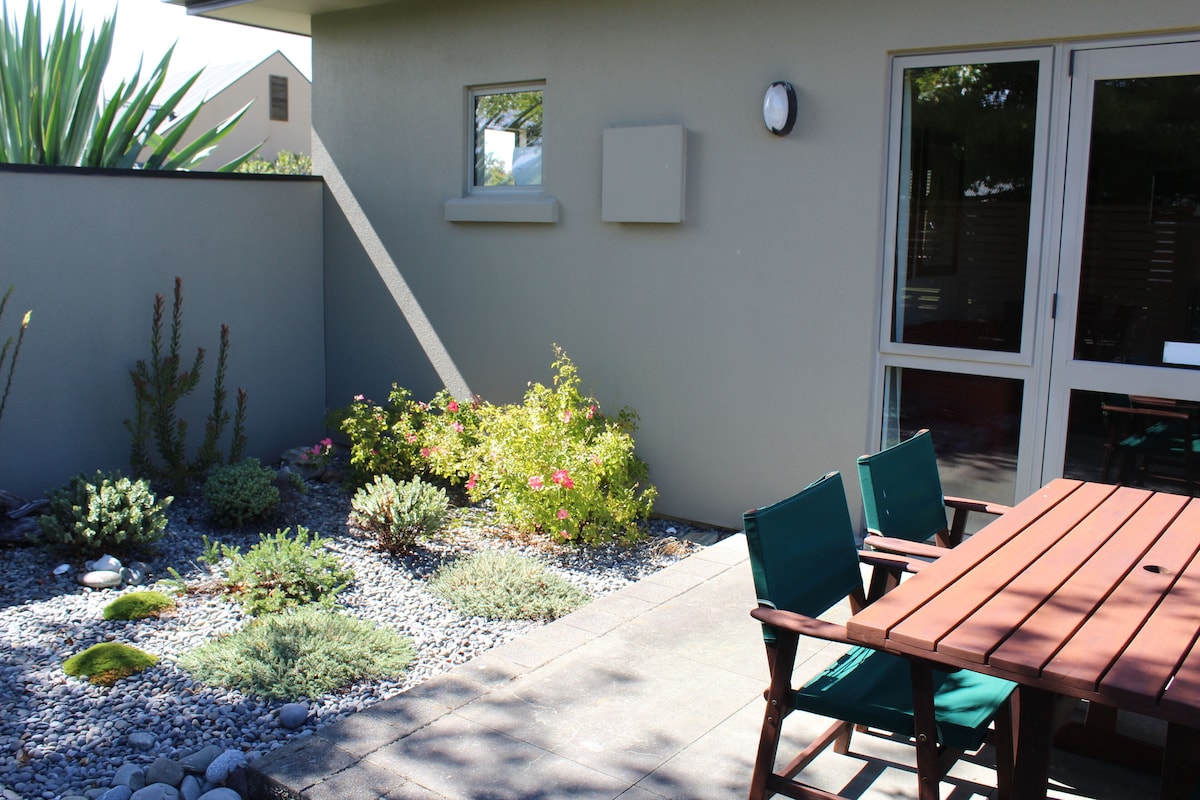 A patio with a wooden table and chairs, surrounded by a garden with various plants and a small window.