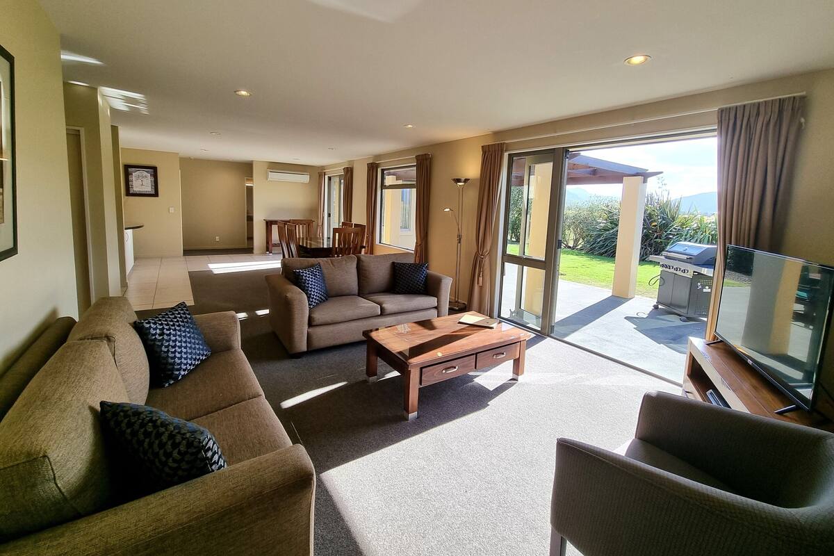 A spacious living room with a sofa set, a coffee table, and a television. There is a sliding glass door leading to a balcony with a barbecue grill.