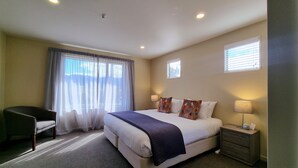 A hotel room with a large bed, a desk, a chair, a nightstand with a lamp, and two windows with blinds. - Vintner's Retreat (Rapaura)