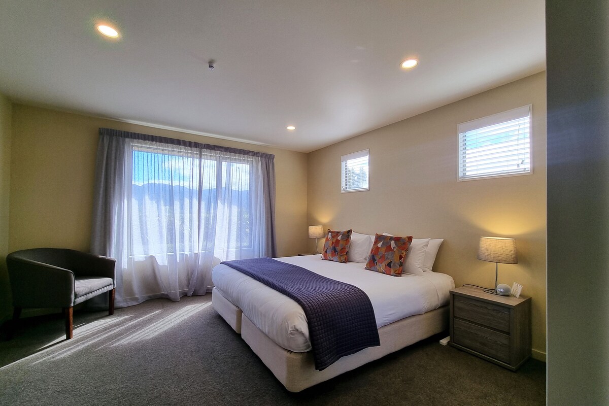 Une chambre d’hôtel avec un grand lit, un bureau, une chaise, une table de chevet avec une lampe et deux fenêtres équipées de stores.