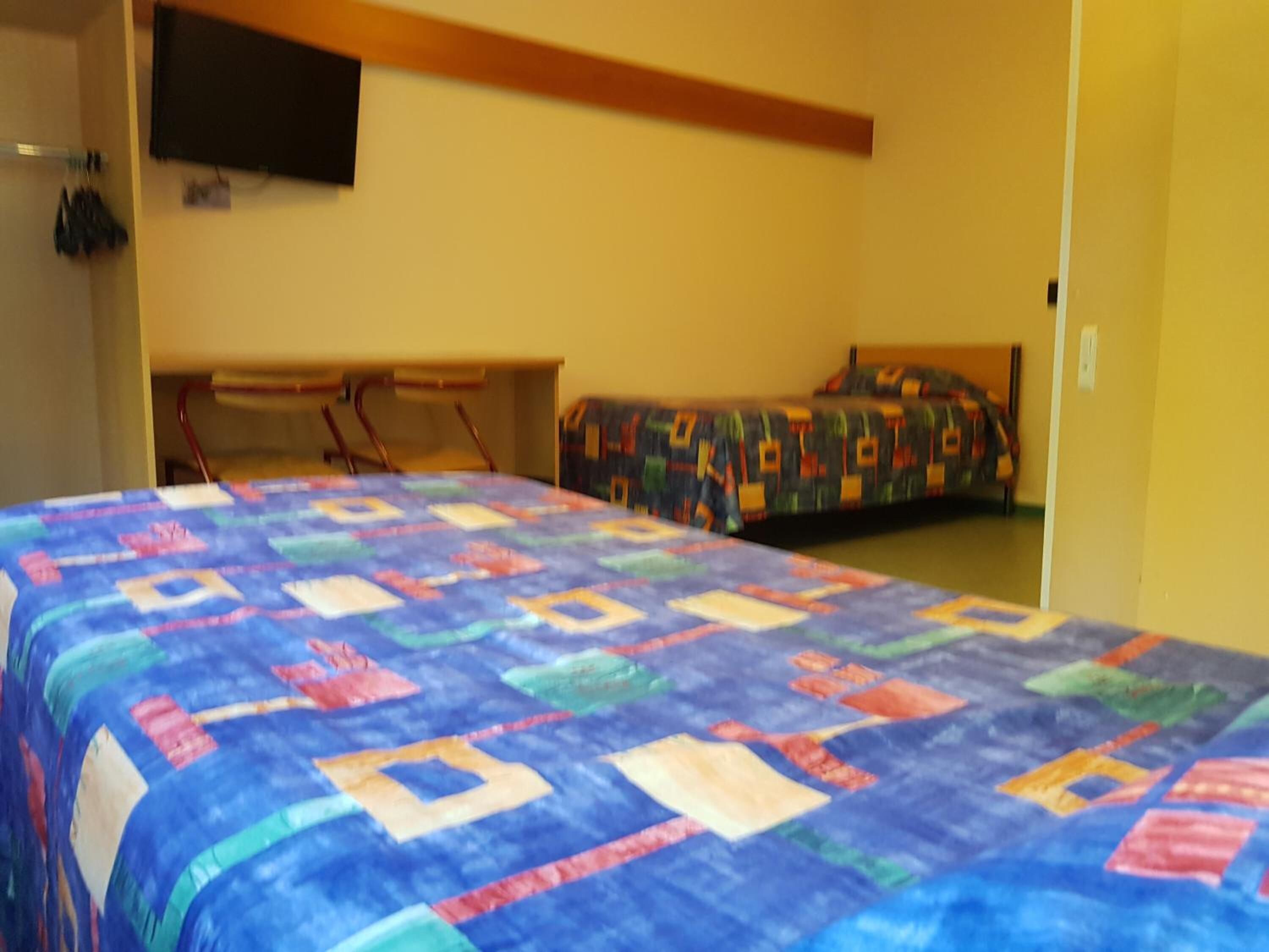 Una habitación con una cama grande, una cama más pequeña, un televisor montado en la pared y una silla.