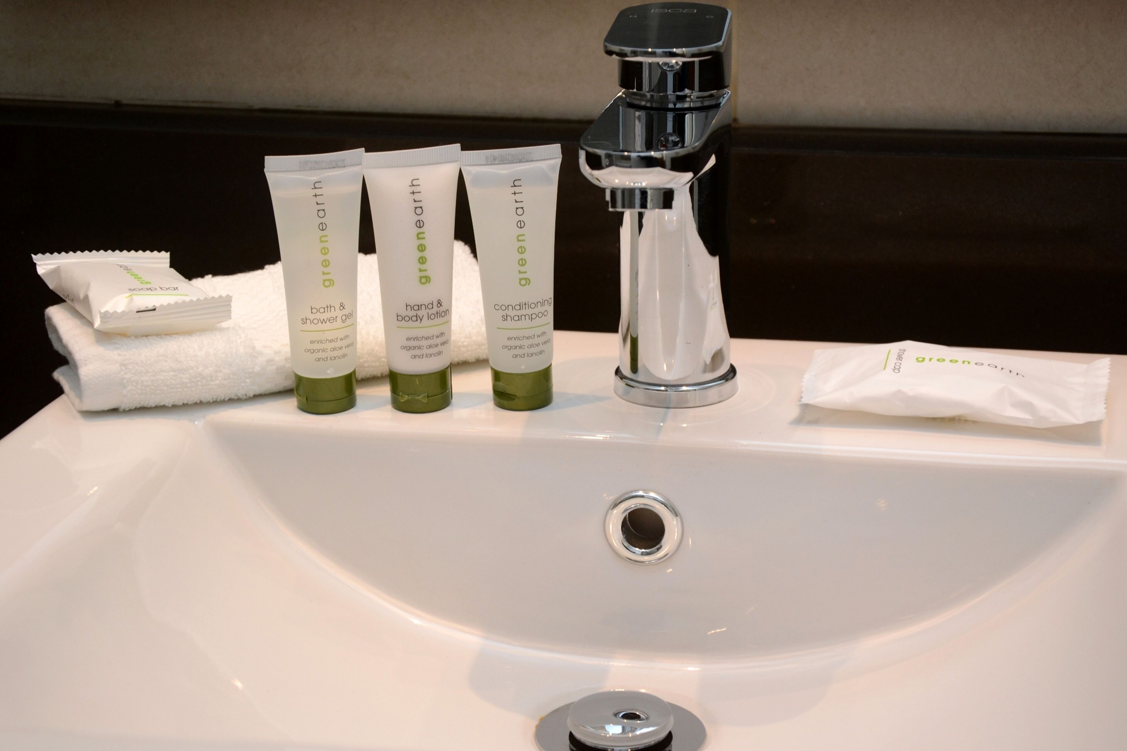 A bathroom sink with three green bottles of Greenearth bath and body products, a white towel, and a faucet.