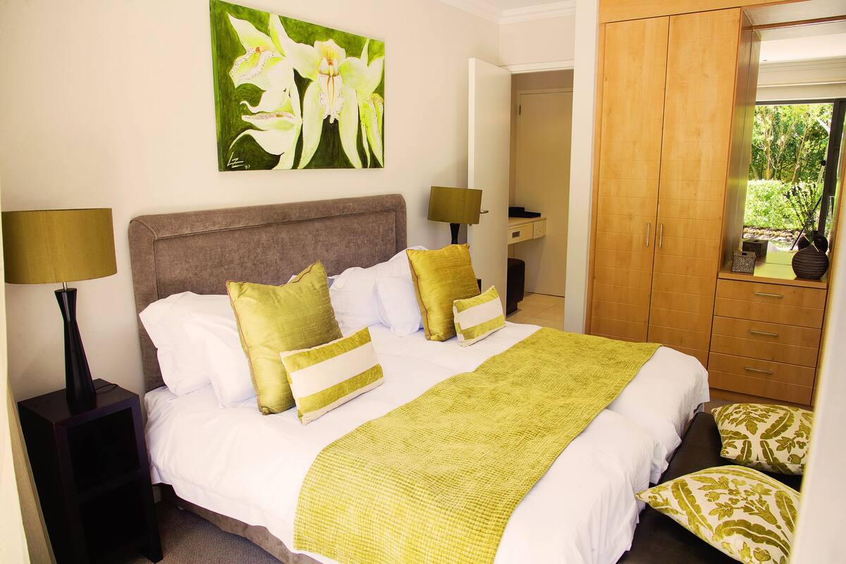 A neatly made bed with a green blanket, white pillows, and a green and white floral painting on the wall.