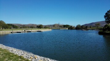 Un lago tranquilo con un paseo de piedra a lo largo de la orilla, rodeado de árboles y colinas al fondo.
