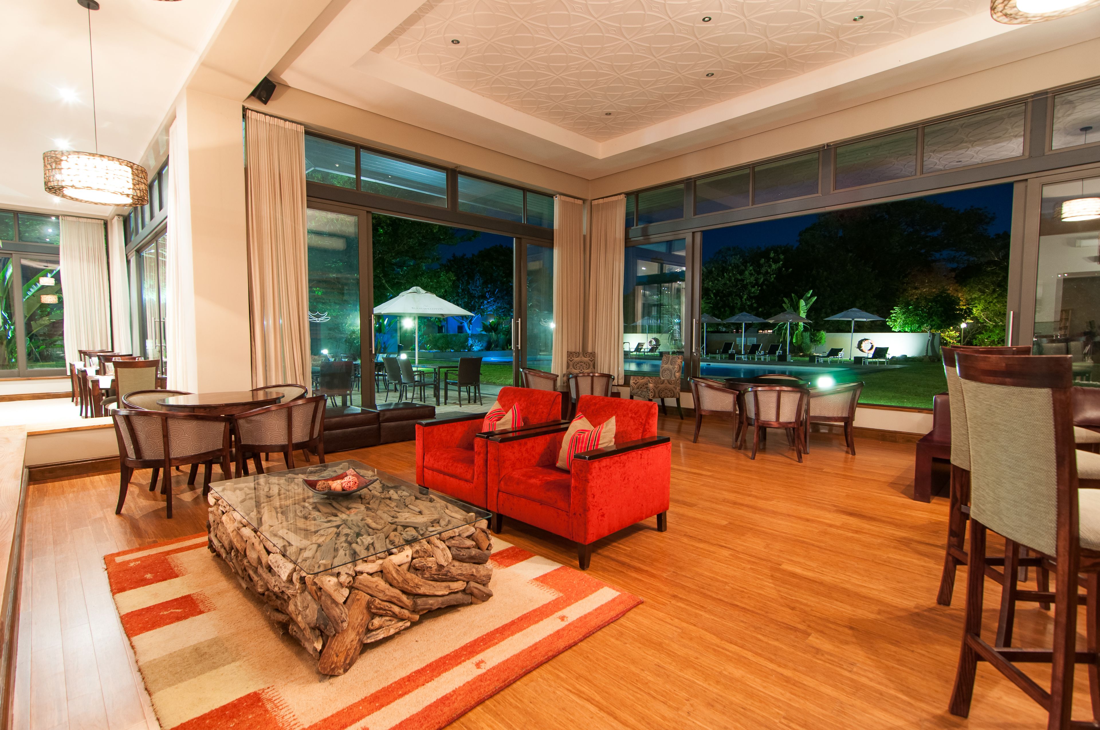 A spacious living area with a red sofa, wooden coffee table, and a dining table with chairs. Large windows offer views of an outdoor pool area.