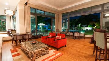 A spacious living area with a red sofa, wooden coffee table, and a dining table with chairs. Large windows offer views of an outdoor pool area.