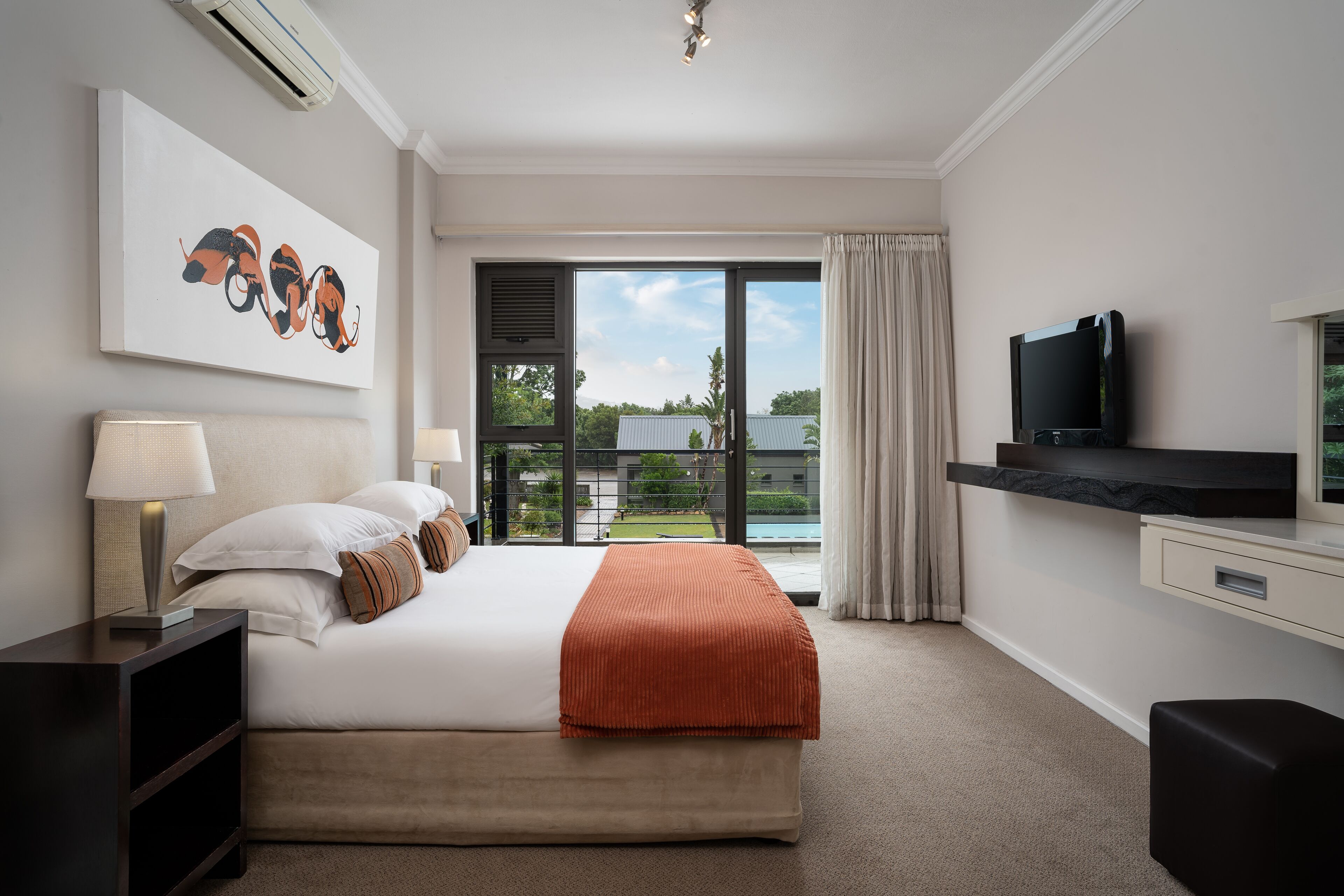 A modern hotel room with a large bed, a flat-screen TV, a desk, and a balcony with a view of the outdoors.