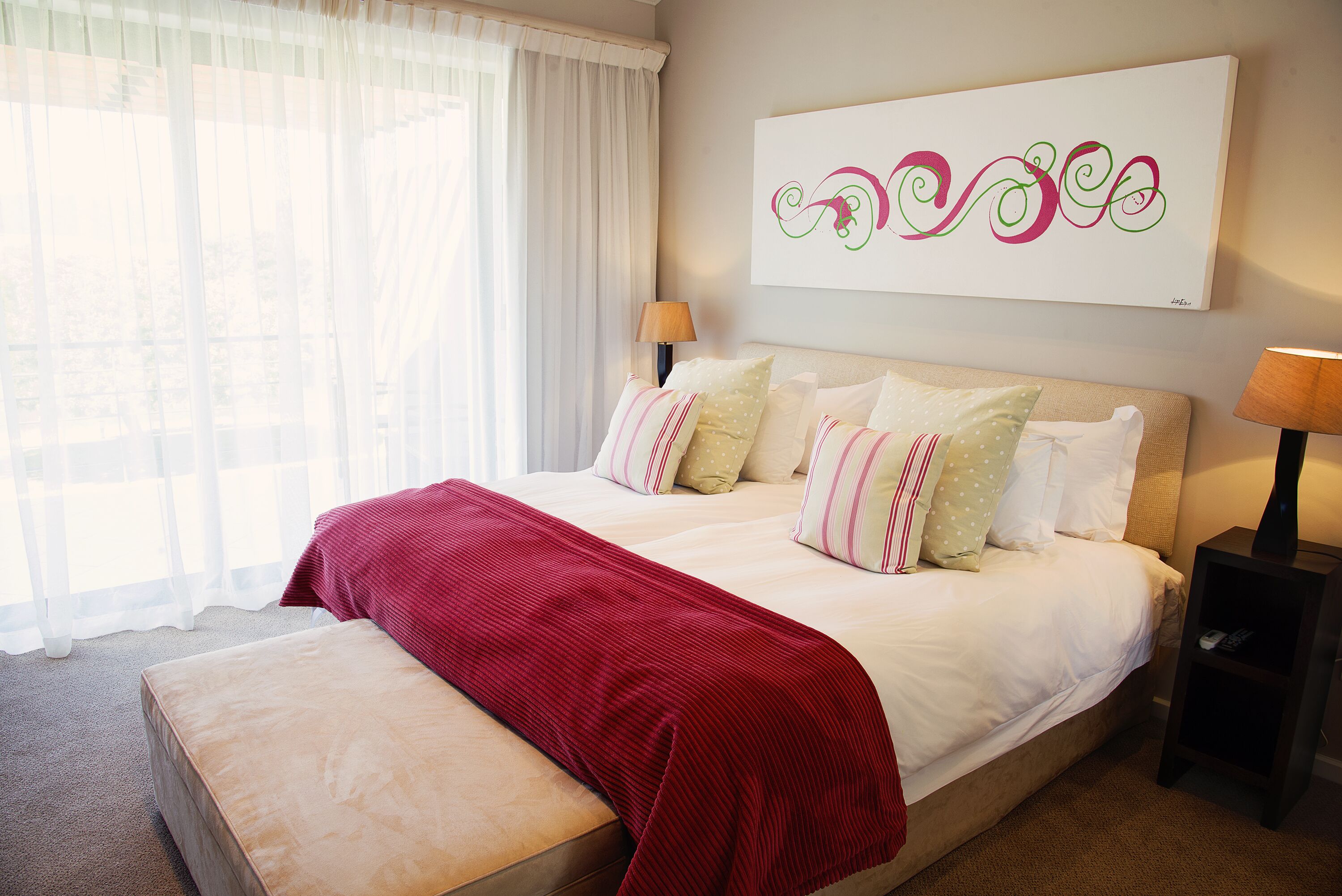 Una cama bien tendida con una manta roja, una mesita de noche con una lámpara y una obra de arte colgada en la pared.