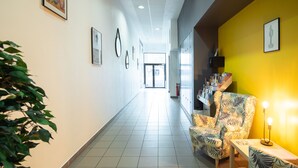 Um corredor de hotel com uma poltrona estampada e uma mesinha, um extintor de incêndio e quadros nas paredes.