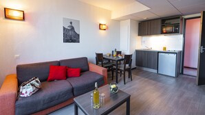 Sala moderna com sofá cinzento, almofada vermelha e uma pequena mesa com garrafa e copos. É visível uma área de jantar com cadeiras e uma cozinha com micro-ondas e pia.