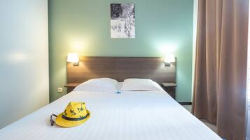 A neatly made bed with white linens, two bedside lamps, a wall-mounted picture, and a yellow hat on the bed.