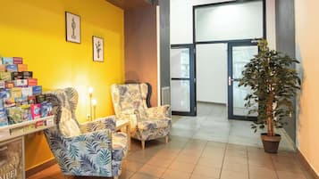 Un espace d’accueil avec un mur jaune, deux fauteuils à motifs, une petite table avec une lampe et une plante en pot.