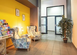 Un espace d’accueil avec un mur jaune, deux fauteuils à motifs, une petite table avec une lampe et une plante en pot.