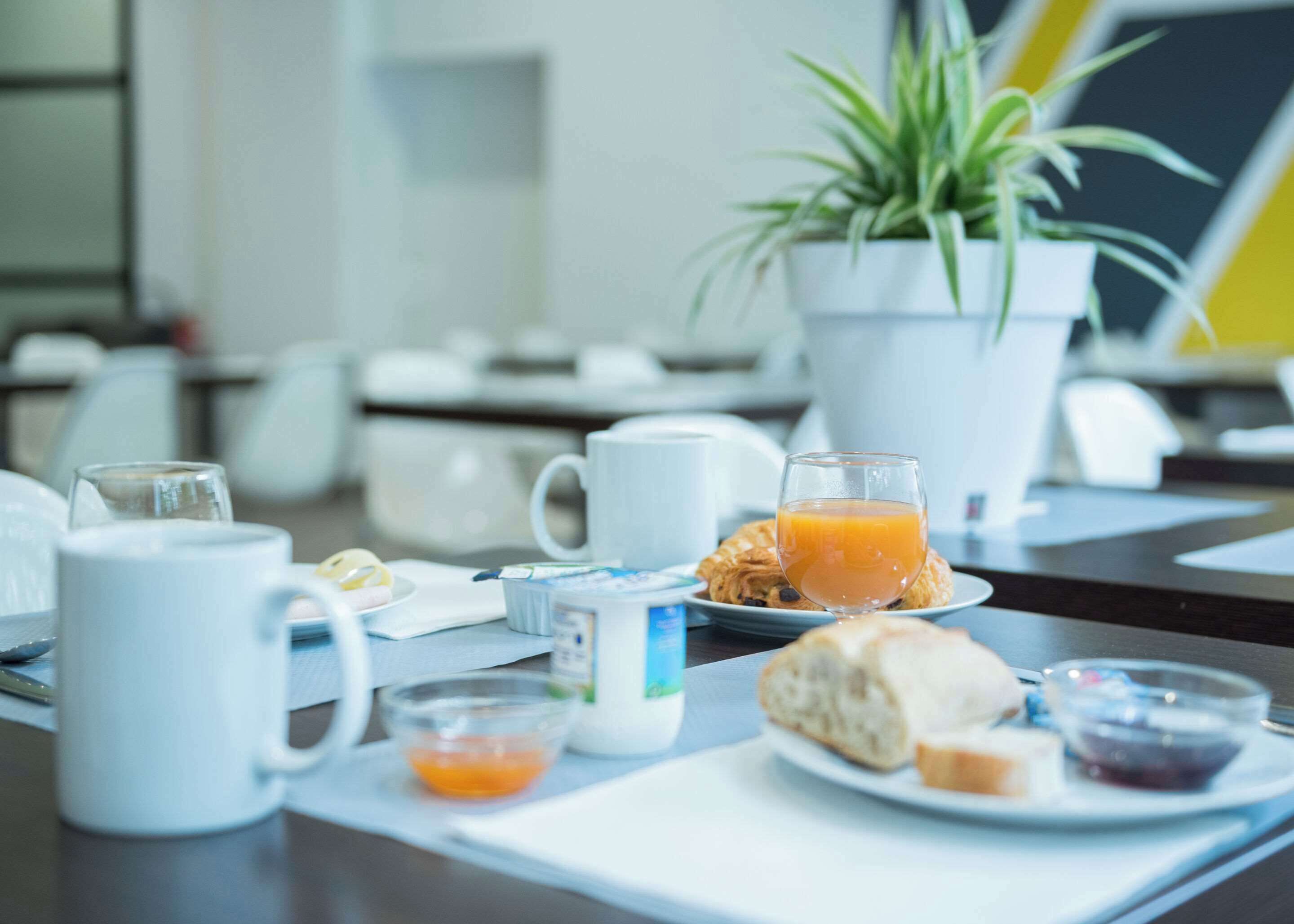 A breakfast setup with a coffee pot, mug, glass of orange juice, croissant, and small bowls of jam and cream.