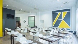 Uma sala de aula moderna com cadeiras e mesas brancas, um relógio e uma grande pintura abstrata na parede.