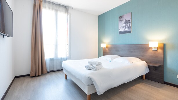 Quarto de hotel com cama, mesa de cabeceira, televisão fixada na parede e janela com cortinas.