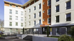Ein mehrstöckiges Hotel mit modernem Design, das eine Kombination aus weißen und braunen Fassaden, großen Fenstern und einem sichtbaren Eingang mit Stufen zum Erdgeschoss zeigt.