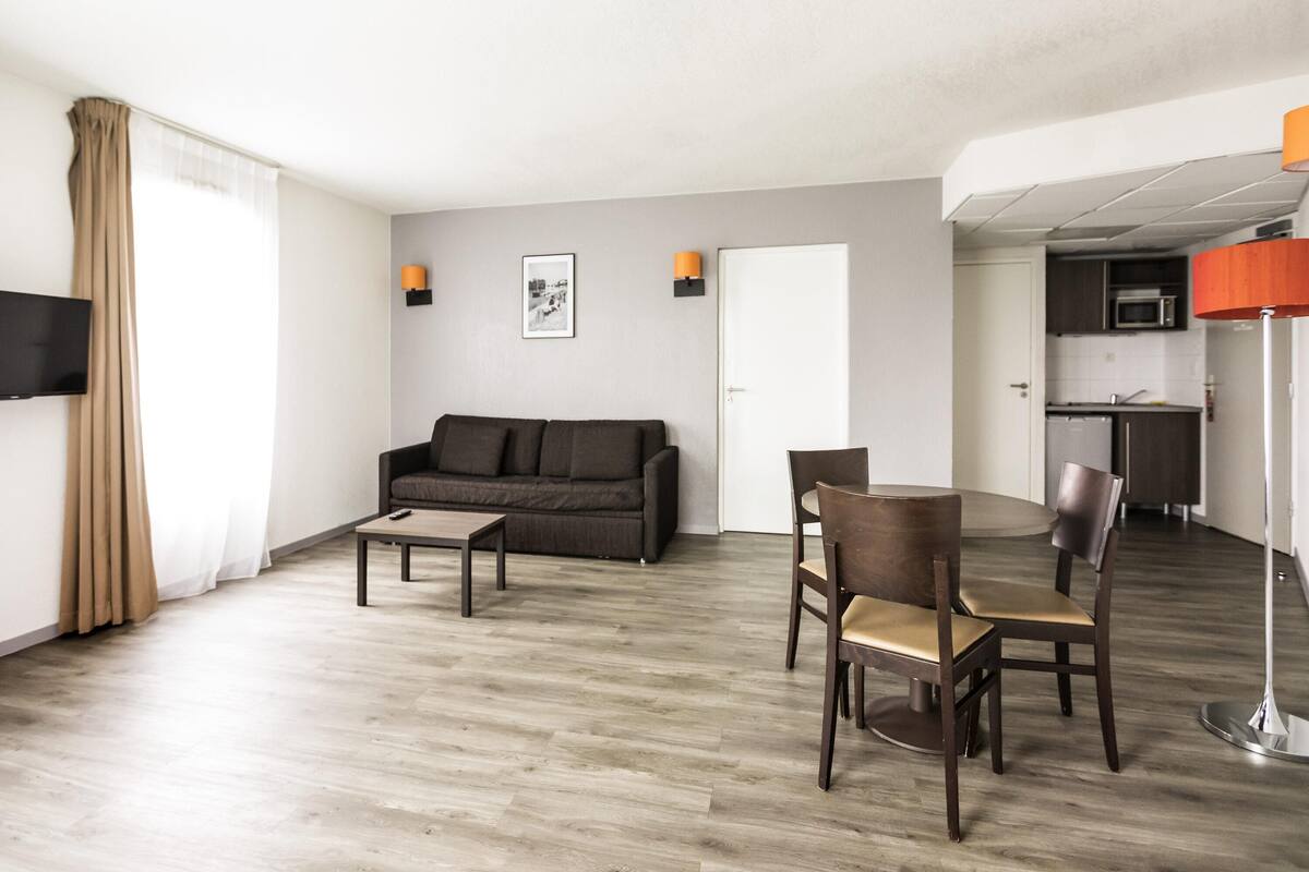 Sala de estar com sofá, mesa de centro, cadeiras, televisão e uma área de cozinha com micro-ondas.