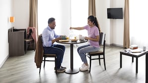 Ein Hotelzimmer mit einem runden Esstisch, zwei Stühlen, einem an der Wand befestigten Fernseher und einem kleinen Tisch mit einem Buch und einer Tasse.