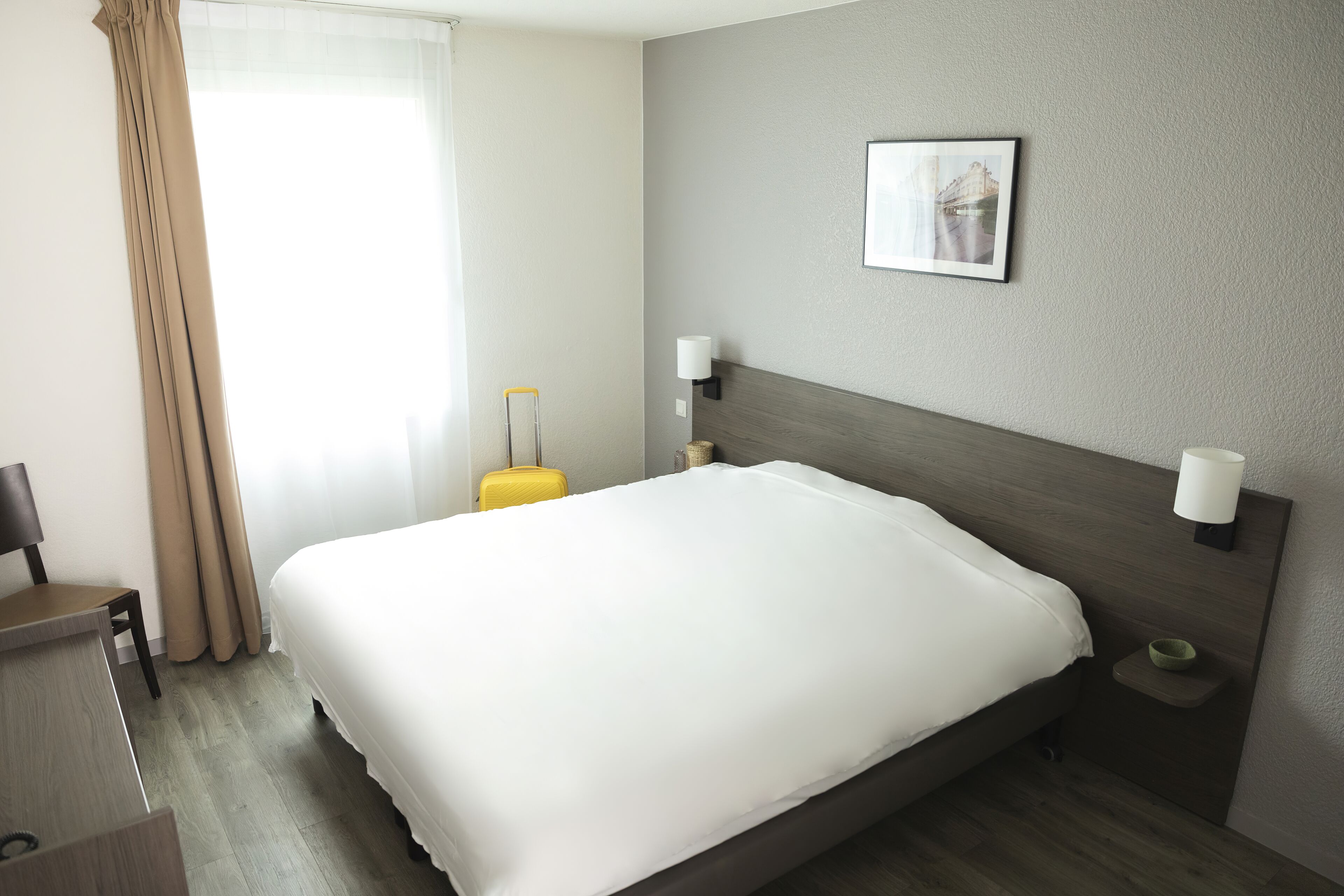 A hotel room with a large bed, a desk, a chair, a suitcase, a wall-mounted lamp, and a picture on the wall.