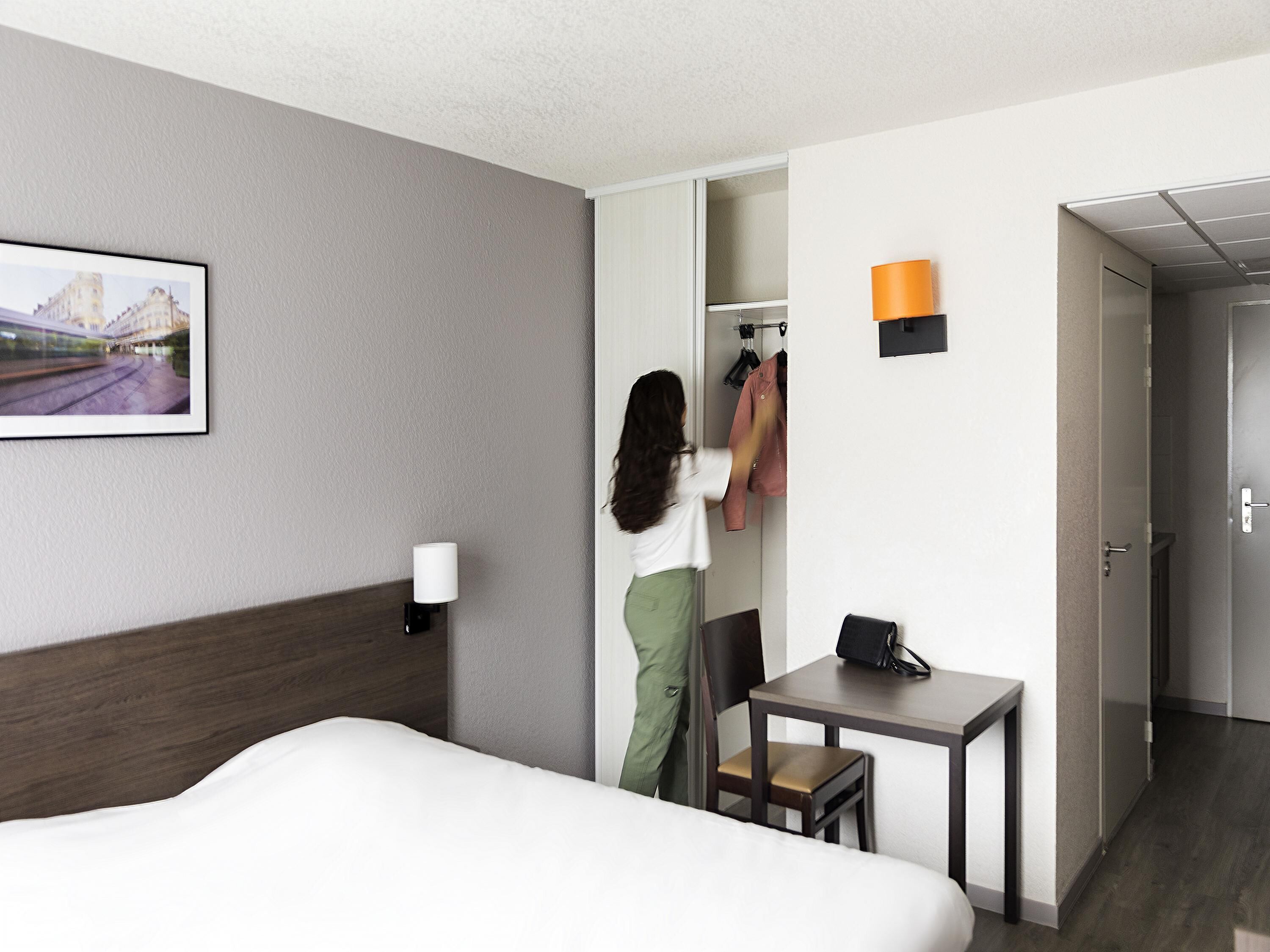 A hotel room with a bed, a desk, a chair, a coat rack, and a picture on the wall.