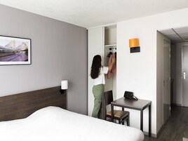 A hotel room with a bed, a desk, a chair, a coat rack, and a picture on the wall.