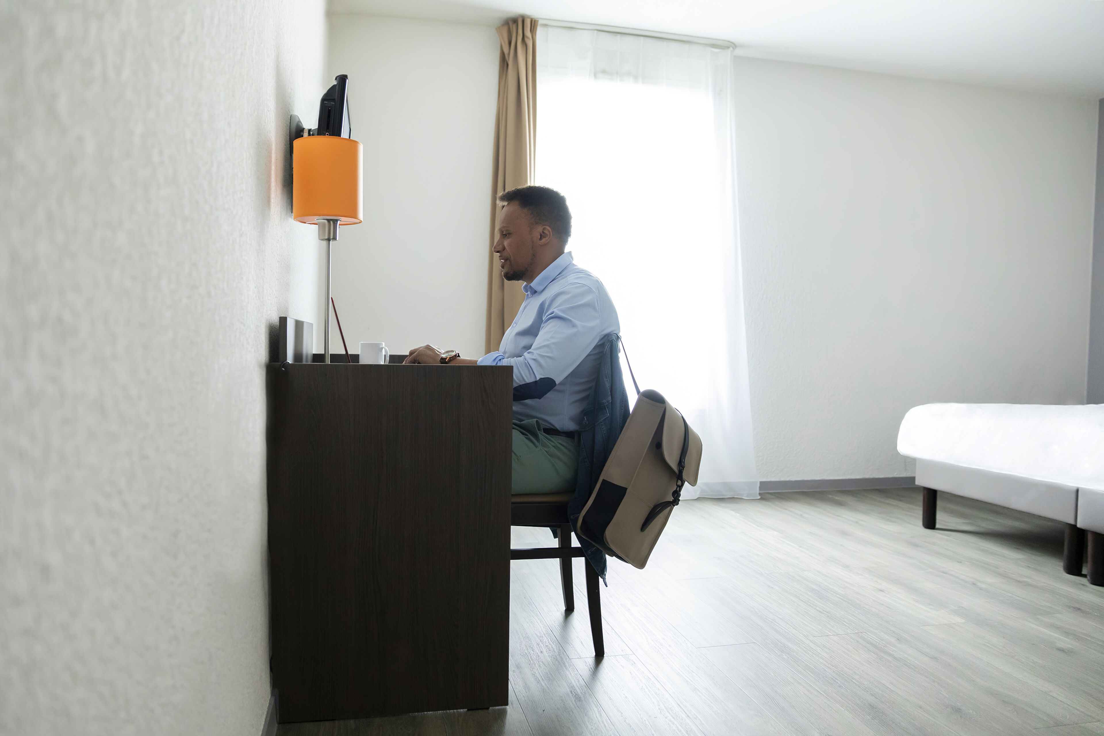 Un hombre sentado en un escritorio en una habitación de hotel, con una cama y una ventana con cortinas.