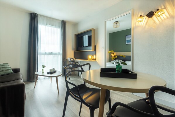 Una sala de estar moderna con un sofá, una mesa de comedor redonda y sillas. Un televisor de pantalla plana está montado en la pared, y hay una mesita auxiliar con un florero y una bandeja.