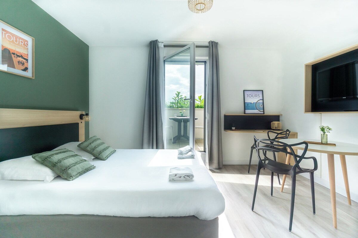 A modern hotel room with a bed, a desk, a chair, a TV, and a balcony with curtains.