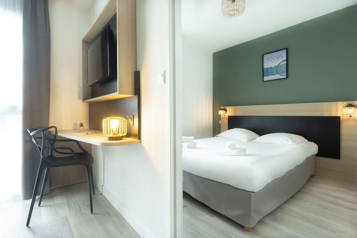 Una habitación de hotel con cama, escritorio con silla, televisor e iluminación empotrada en la pared.