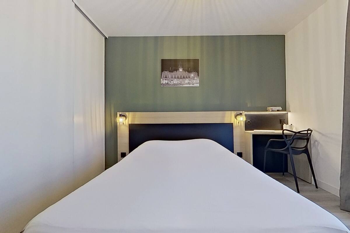Una habitación de hotel con cama, escritorio con silla, un cuadro en la pared y una mesita de noche con lámpara.