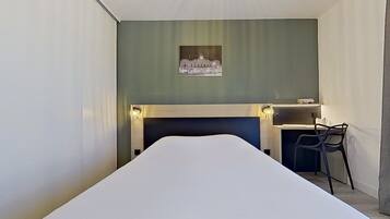 A hotel room with a bed, a desk with a chair, a wall-mounted picture, and a small table with a lamp.