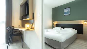 Quarto de hotel com cama, escrivaninha com cadeira, televisão e luminária na parede.