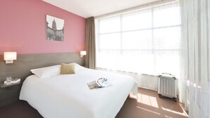 Habitación de hotel con una cama grande, una ventana con cortinas translúcidas, un radiador y una mesita de noche con una lámpara.