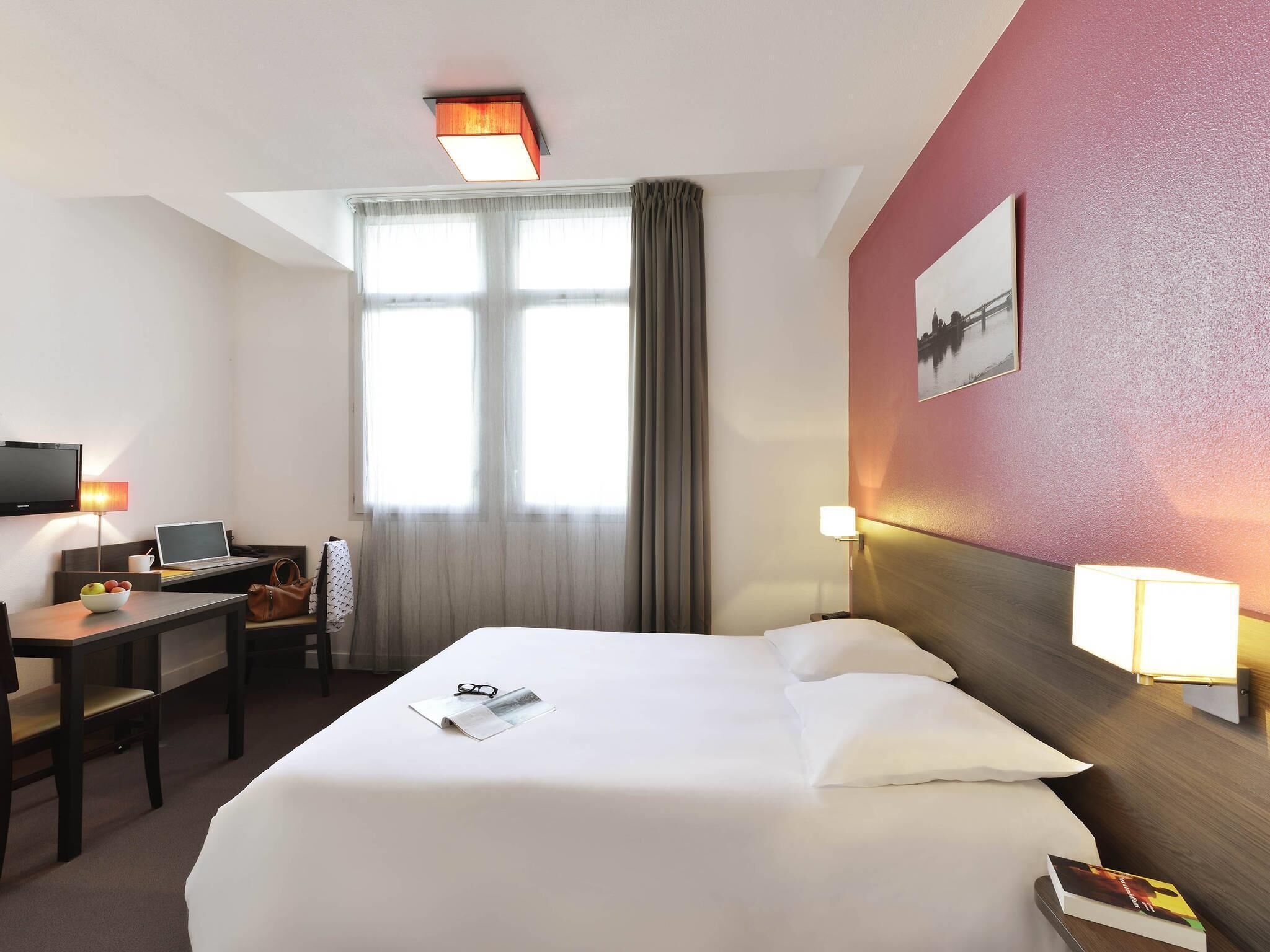 A hotel room with a bed, desk, chair, lamp, and a window with curtains.