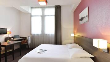 A hotel room with a bed, desk, chair, lamp, and a window with curtains.