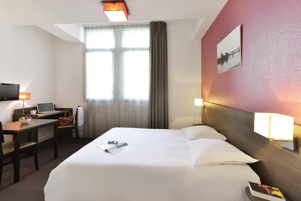 Habitación de hotel con cama, escritorio, silla, televisor y ventana con cortinas.