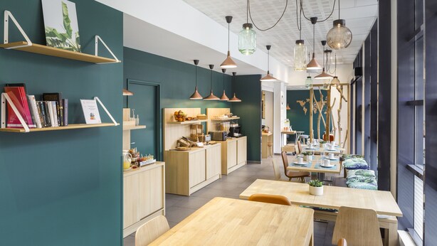 Un comedor moderno y bien iluminado con mesas y sillas de madera, una zona de cocina con armarios de madera e iluminación colgante.