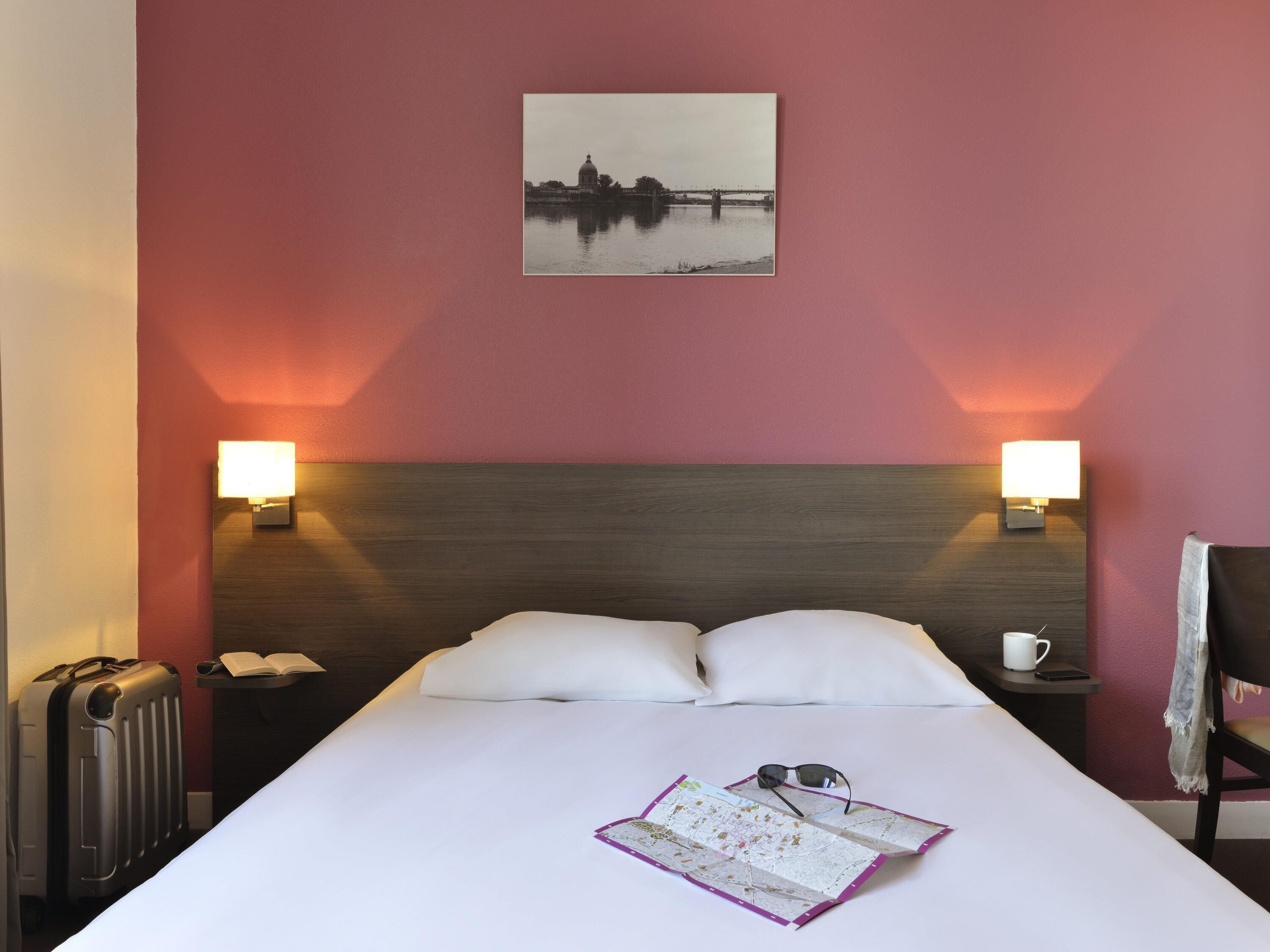 A hotel room with a large bed, two bedside lamps, a map, a pair of sunglasses, and a suitcase.