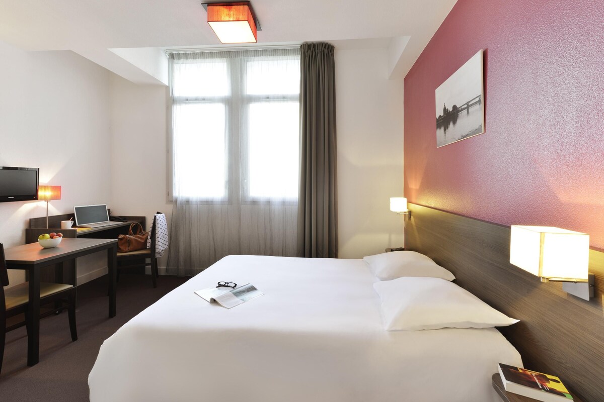 Habitación de hotel con cama, escritorio, silla, televisor y ventana con cortinas.